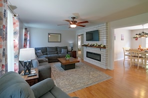 A living room with a sectional sofa, a coffee table, a fireplace, a TV mounted on a brick wall, and a dining area with a table and chairs. - Private Yard: Fire Pit & Cornhole! Moorefield Gem (Moorefield)