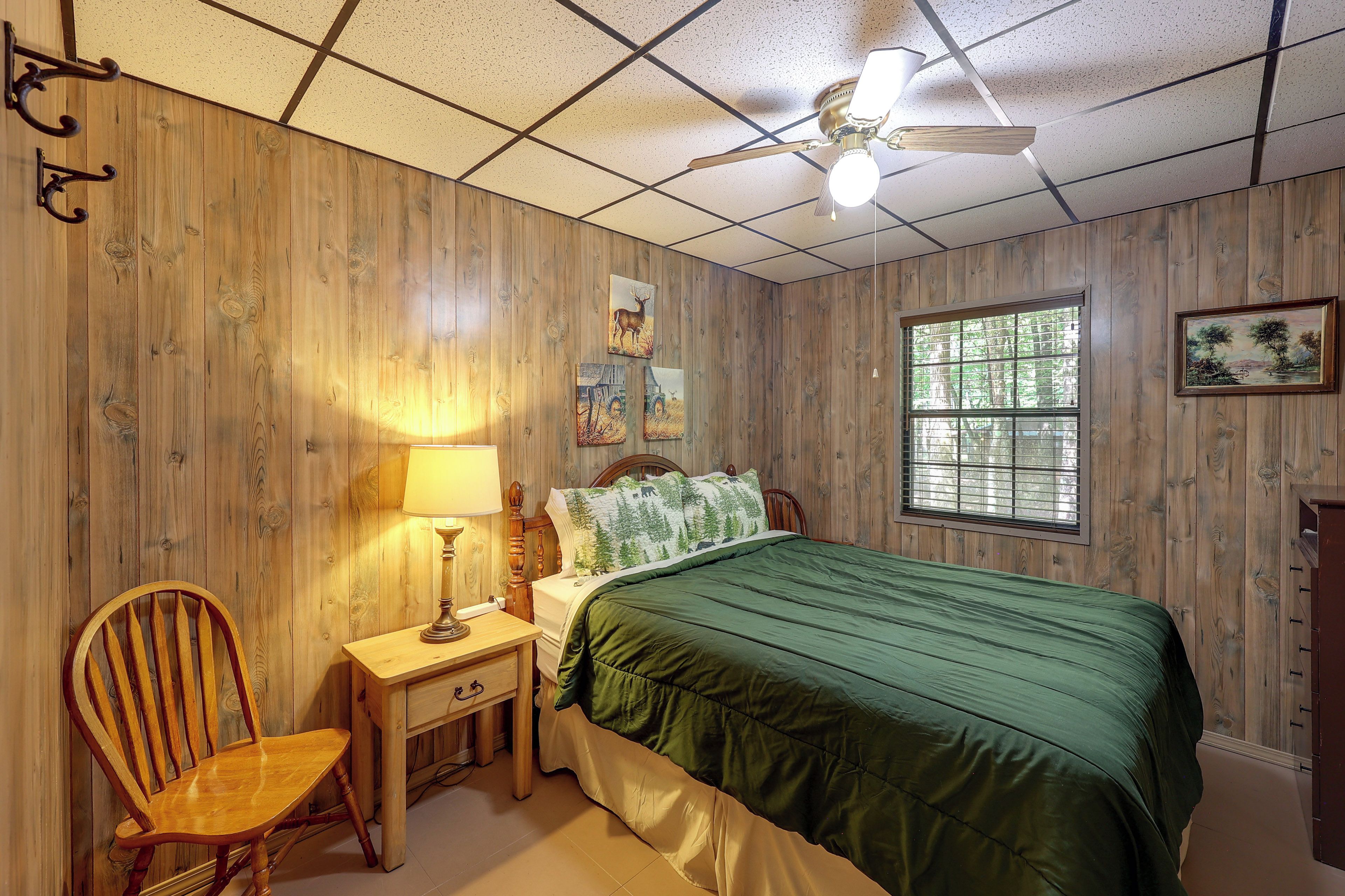 A room with wooden paneling, a bed with a green bedspread, a nightstand with a lamp, a chair, and a window with blinds.
