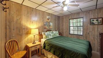 A room with wooden paneling, a bed with a green bedspread, a nightstand with a lamp, a chair, and a window with blinds.