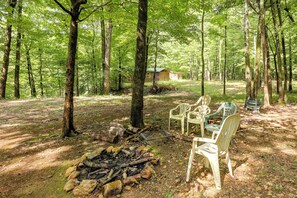 A forested area with a small cabin, outdoor seating, and a fire pit.