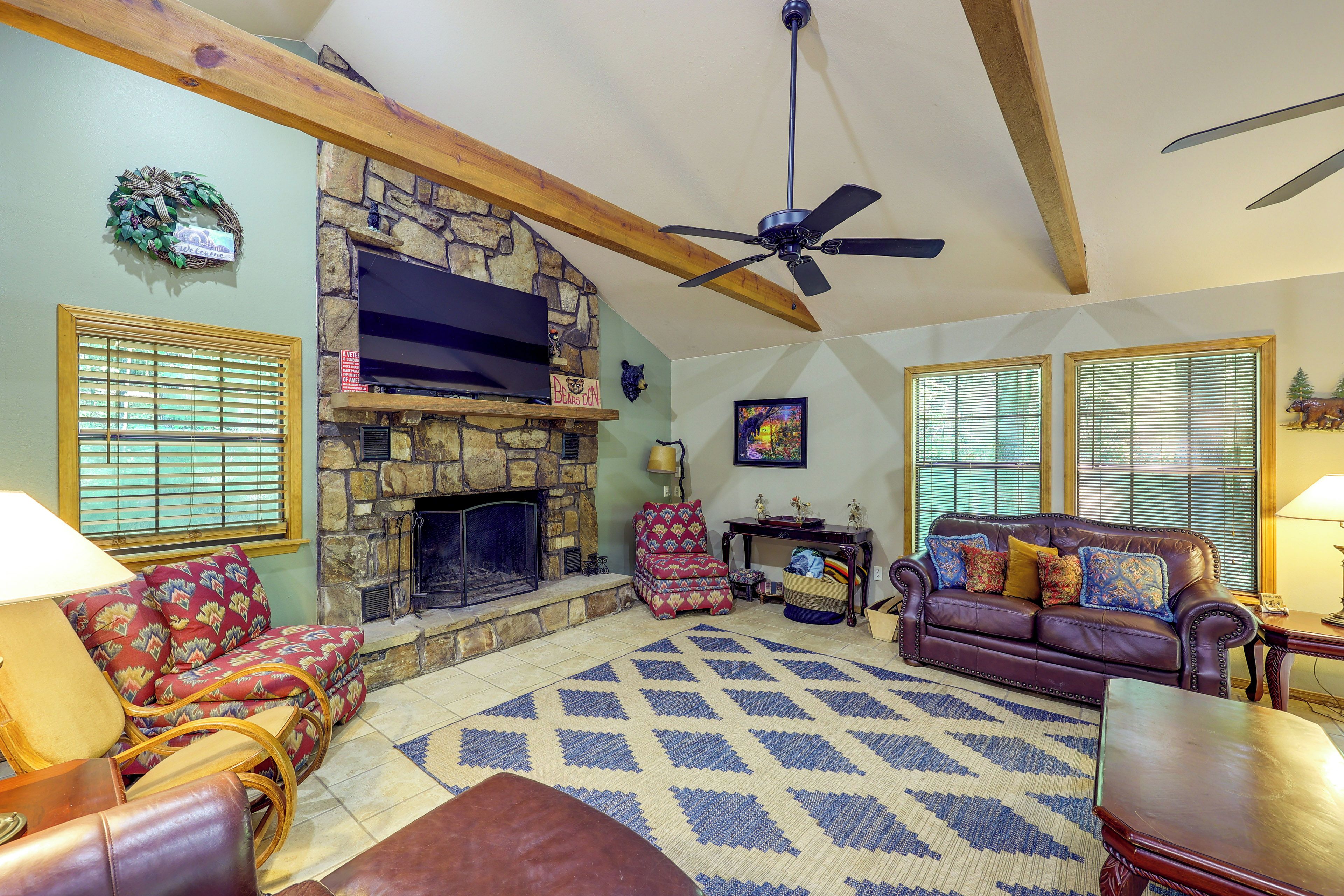 A cozy living room with a stone fireplace, a ceiling fan, and a large TV mounted above it.
