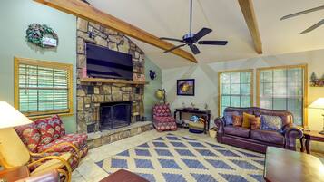 A cozy living room with a stone fireplace, a ceiling fan, and a large TV mounted above it.
