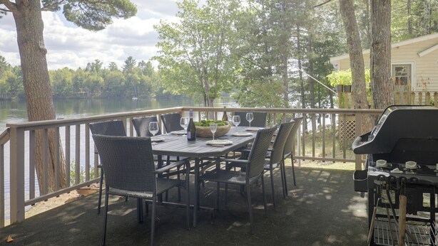 An outdoor dining area with a table set for two, surrounded by chairs, overlooking a body of water with trees in the background. - Lakefront Crystal Falls Home w/ Private Dock (Crystal Falls)