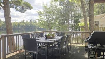 An outdoor dining area with a table set for two, surrounded by chairs, overlooking a body of water with trees in the background.