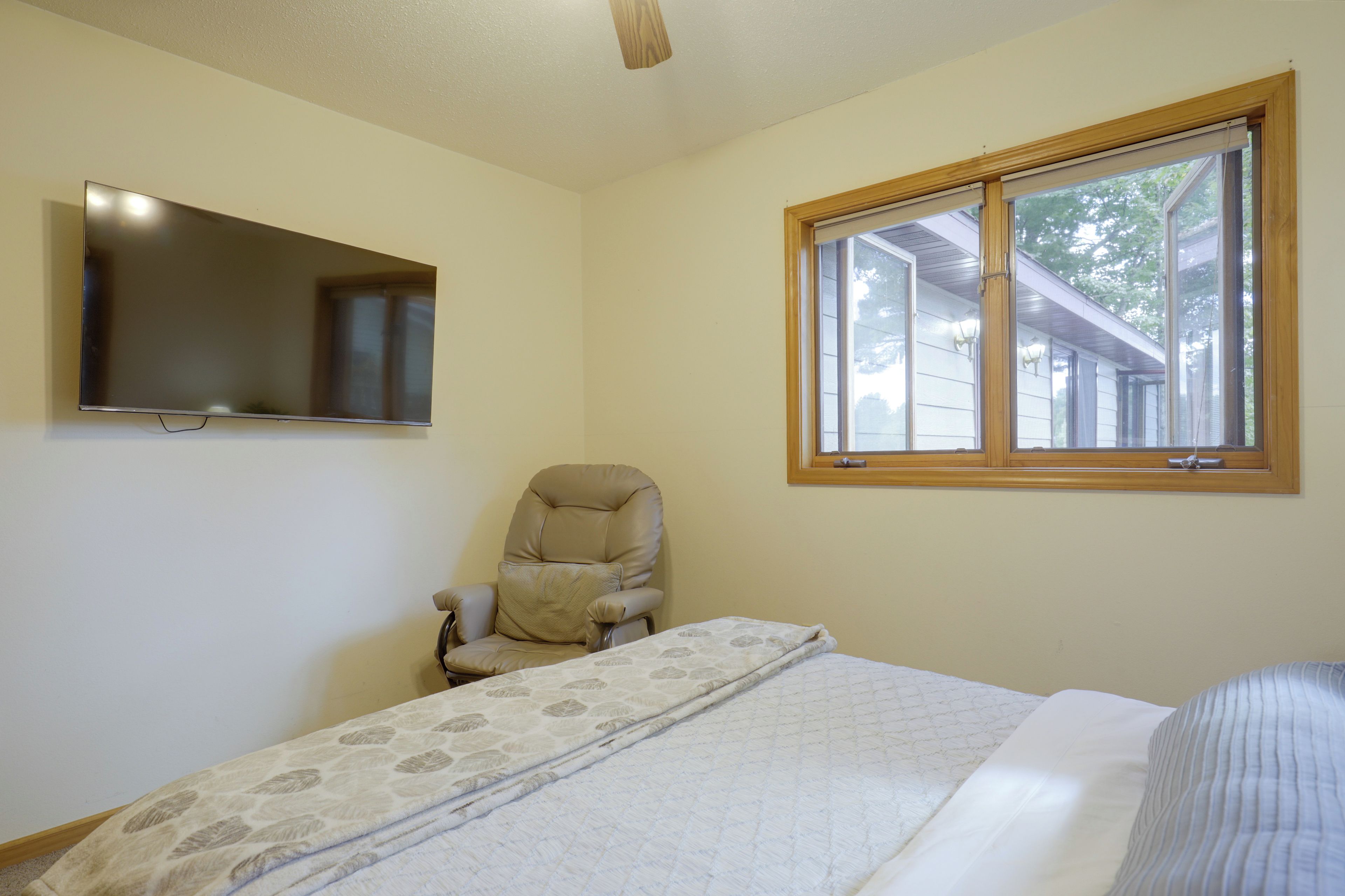 A bedroom with a bed, a chair, a television mounted on the wall, and a window with a view of the outside.