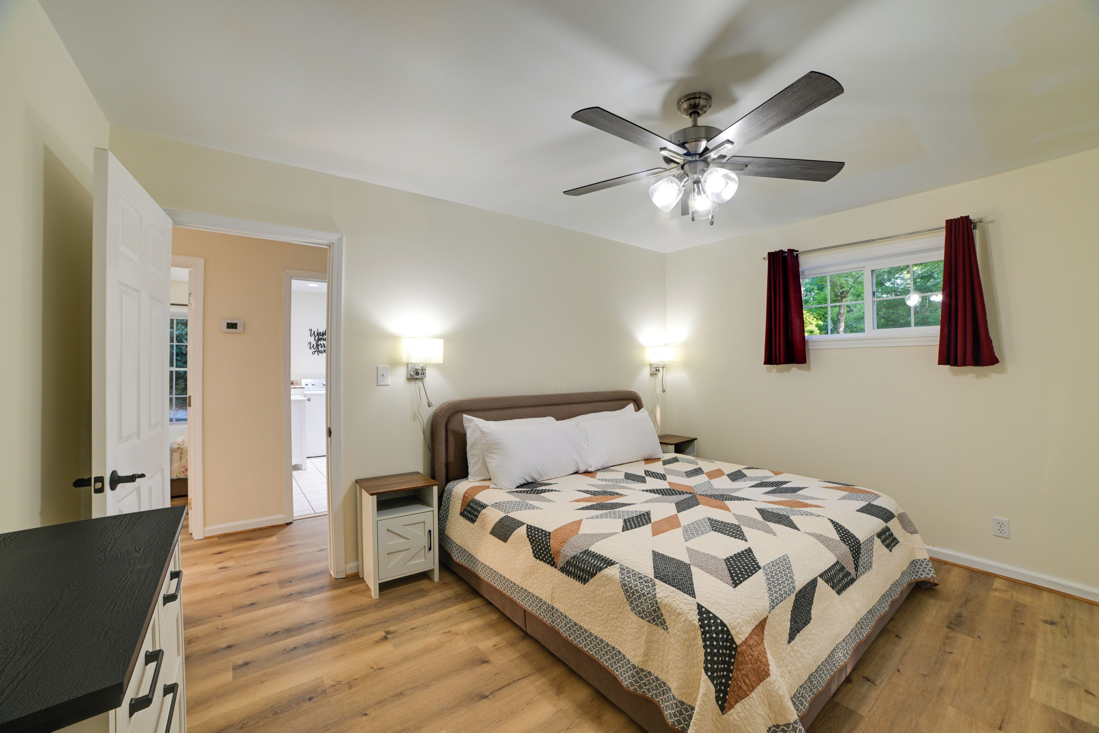 A bedroom with a bed, a ceiling fan, bedside lamps, a window with curtains, and a door leading to another room.