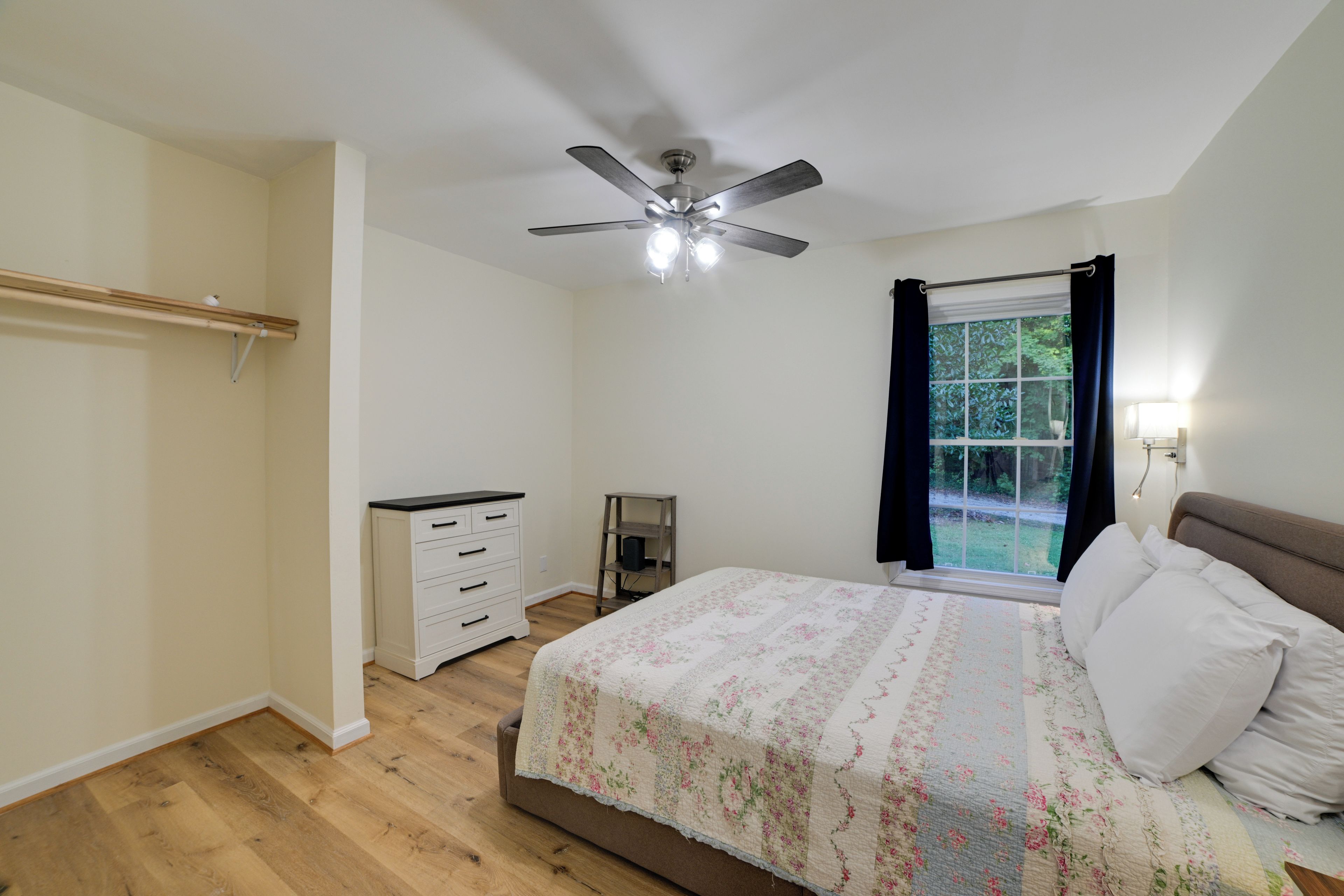 A bedroom with a bed, a ceiling fan, a window with curtains, a bedside table, and a wardrobe.