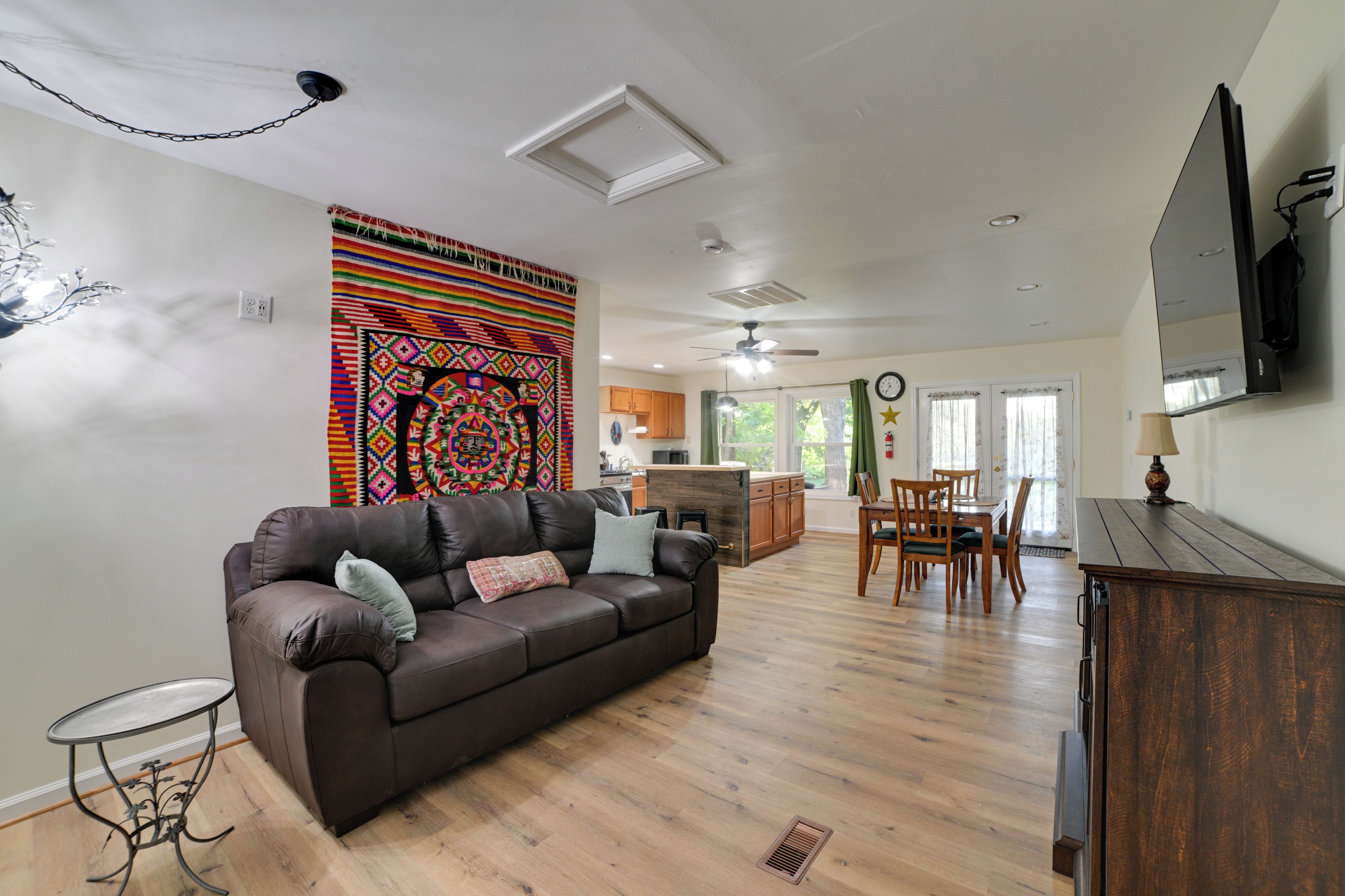 A living room with a brown leather sofa, a dining table with chairs, a kitchen area, and a large colorful tapestry on the wall.