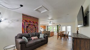 A living room with a brown leather sofa, a dining table with chairs, a kitchen area, and a large colorful tapestry on the wall.