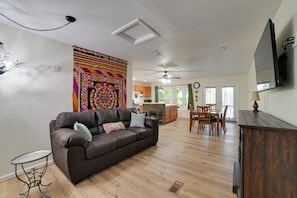 Sala de estar com sofá de couro marrom, mesa de jantar com cadeiras, área de cozinha e uma grande tapeçaria colorida na parede.