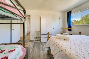 A bedroom with bunk beds, a single bed, a wooden chair, a small stool, and a window with a view of trees.