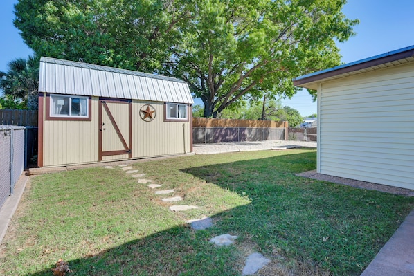 A small, single-story building with a metal roof, a wooden fence, and a grassy yard with stepping stones.