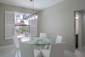 A dining area with a glass table surrounded by chairs, a ceiling light fixture, and a view through a window with shutters. - Brand-new Luxury Home w/ Pool! 9 Mi to Pine Island (Cape Coral)