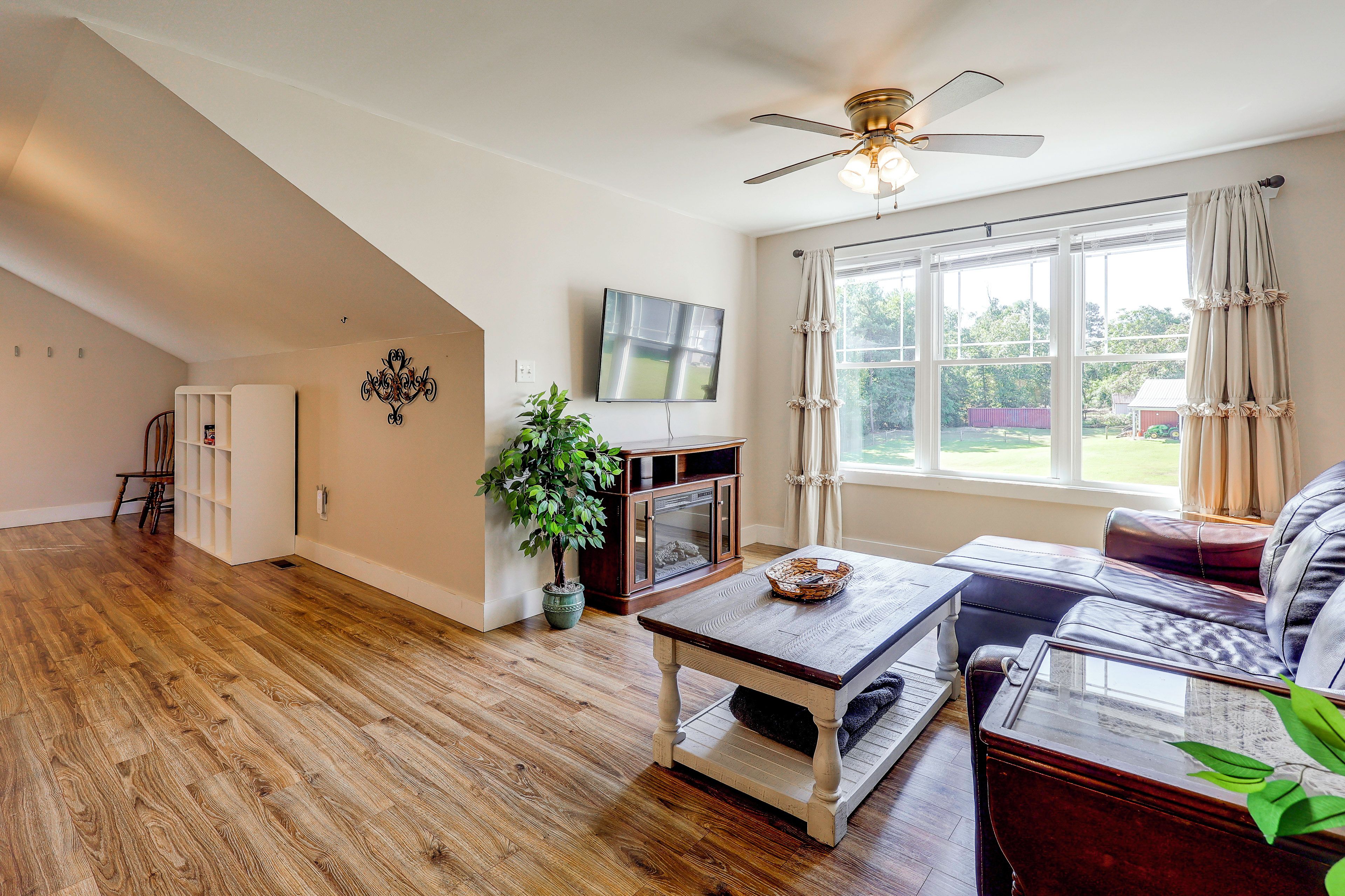 A spacious living room with hardwood flooring, a ceiling fan, a television, and a large window with curtains.