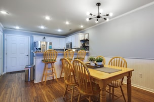Una cocina con armarios blancos, un refrigerador de acero inoxidable, un microondas y un comedor con sillas de madera y una mesa.