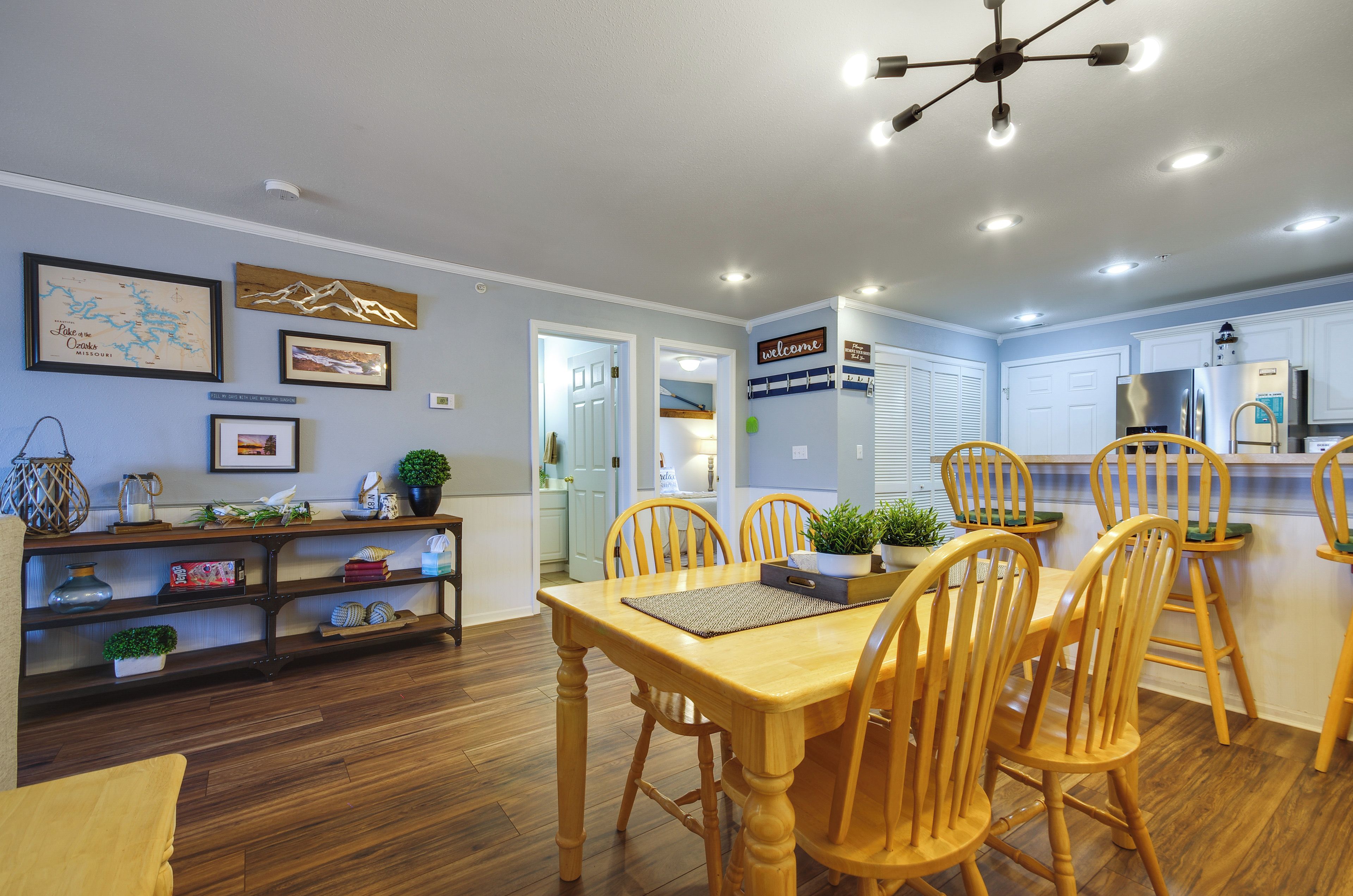A dining area with a wooden table and chairs, a kitchen in the background, and a ceiling light fixture.