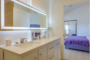 A bathroom with a large mirror, two sinks, a towel rack, and a bed with a purple blanket in the adjacent room. - Extended Stay Near Beaches & LA Commuter Routes (Torrance)