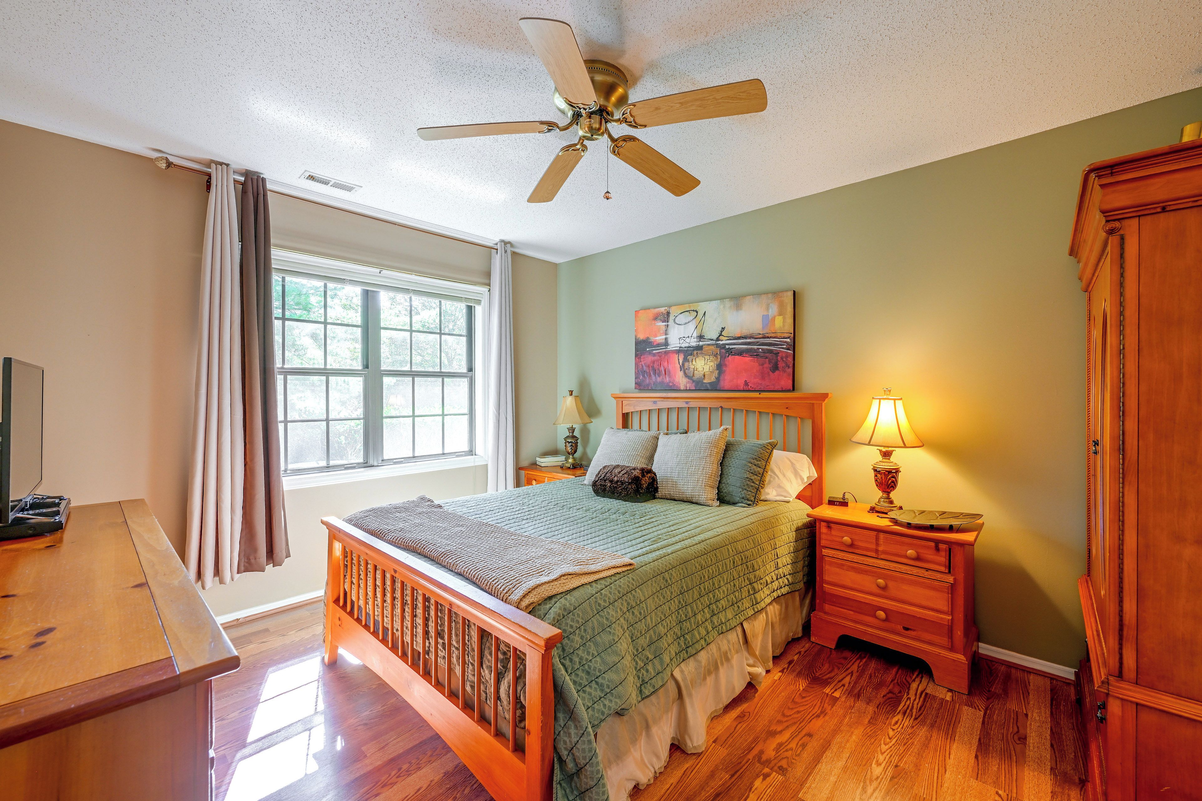 A bedroom with a bed, a nightstand, a lamp, a ceiling fan, a window with curtains, and a painting on the wall.