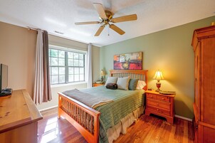 A bedroom with a bed, a nightstand, a lamp, a ceiling fan, a window with curtains, and a painting on the wall.