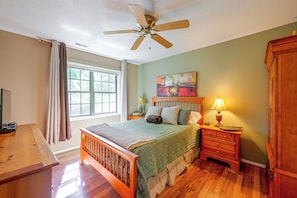 A bedroom with a bed, a nightstand, a lamp, a ceiling fan, a window with curtains, and a painting on the wall.