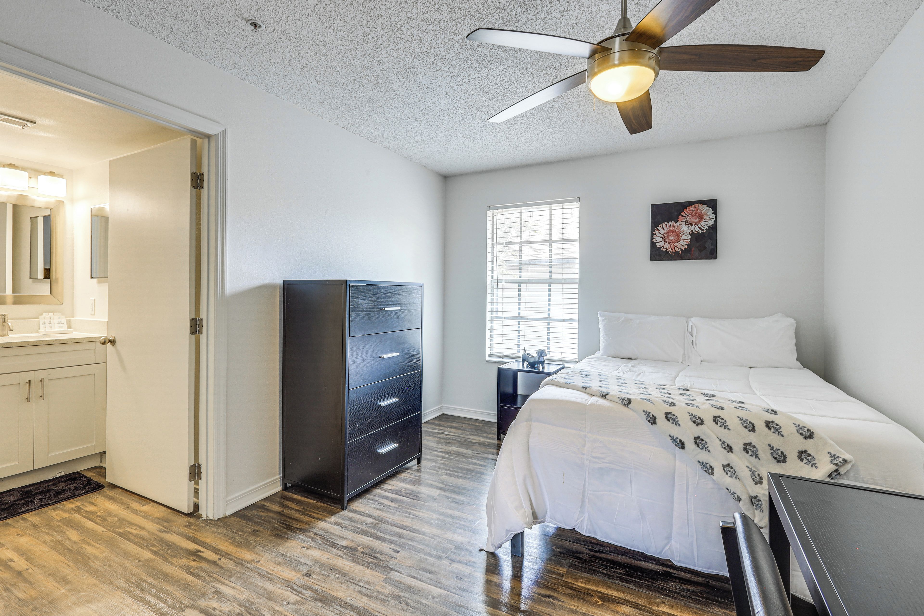 A bedroom with a bed, a dresser, a ceiling fan, a window with blinds, and a wall-mounted artwork.