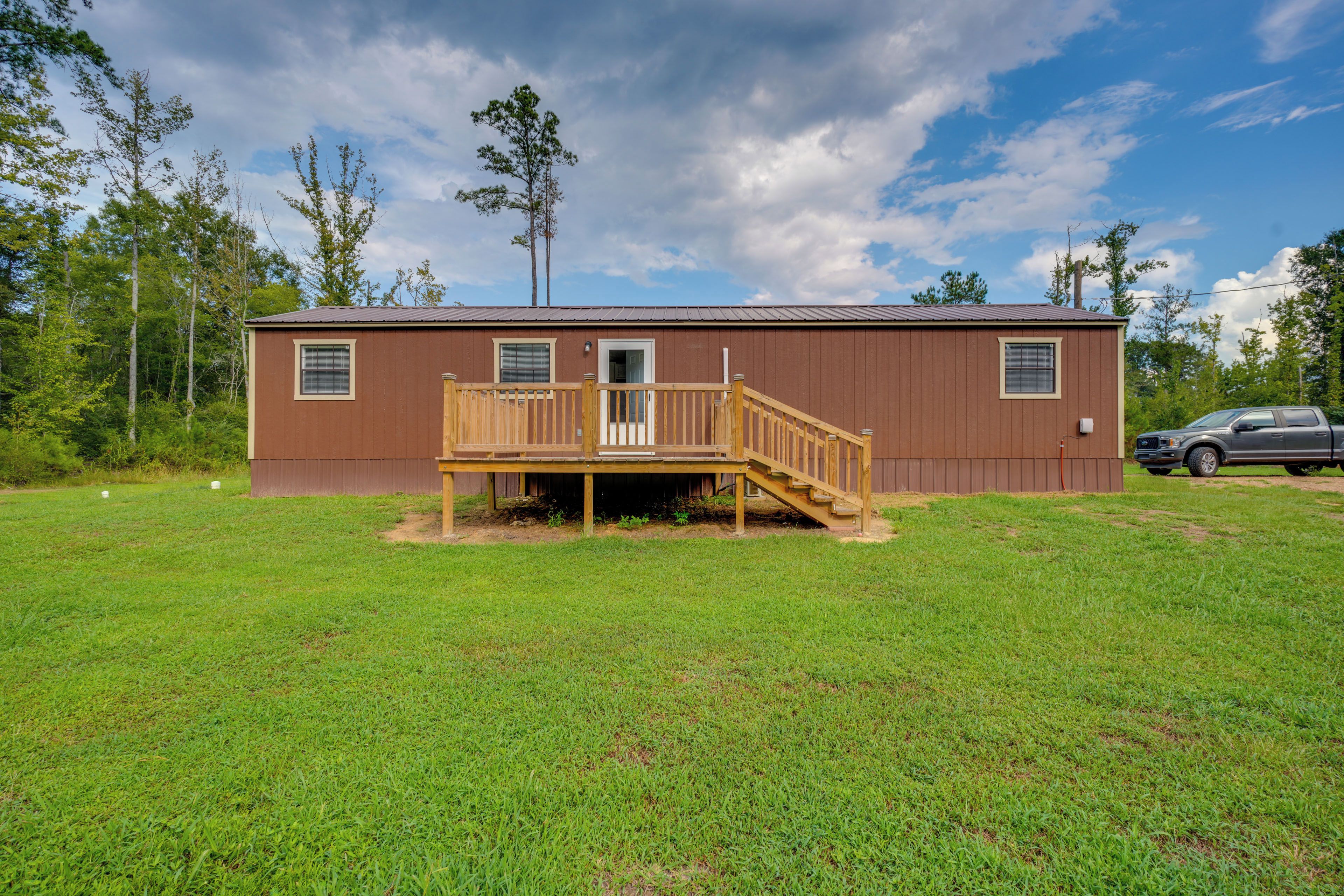 A single-story wooden cabin with a deck, surrounded by a grassy field and trees.