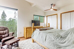 Ein Schlafzimmer mit Bett, einem Sessel, einem Schreibtisch, einem Deckenventilator und einem Fenster mit Blick auf Bäume.