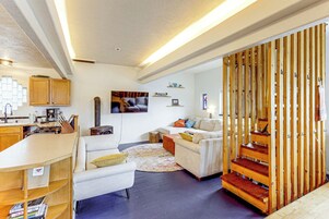 A modern living room with a kitchen area, wooden staircase, and a flat-screen TV mounted on the wall.