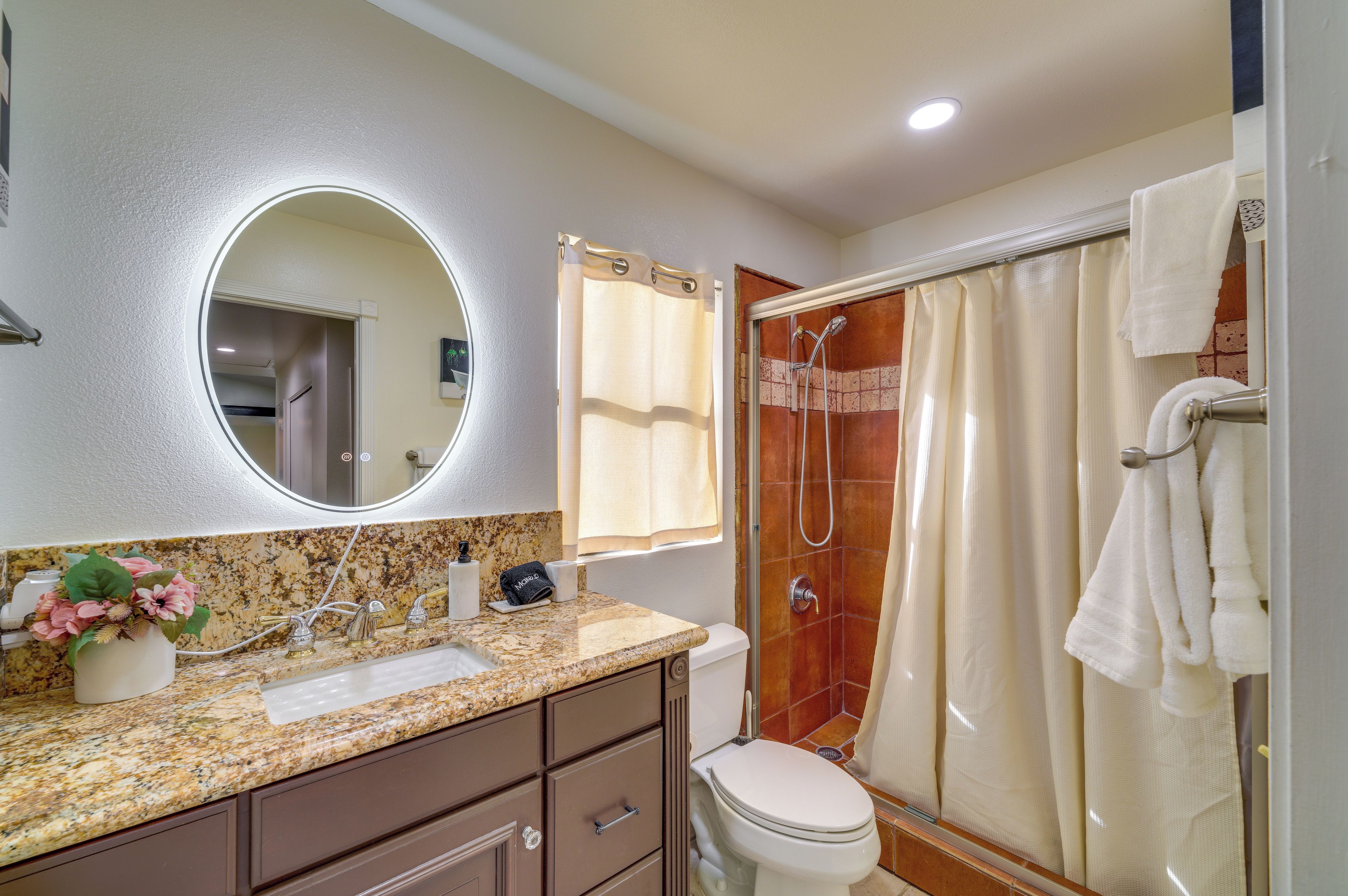 Une salle de bain dotée d’un comptoir en granit, de toilettes, d’une douche avec une porte en verre et d’un miroir à cadre circulaire.