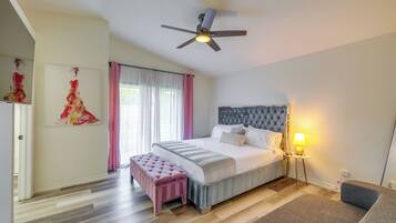 Un dormitorio con cama, mesita de noche con lámpara, ventilador de techo, ventana con cortinas y una obra de arte en la pared.