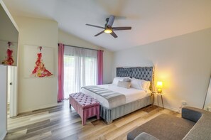 A bedroom with a bed, a bedside table with a lamp, a ceiling fan, a window with curtains, and a wall-mounted artwork.