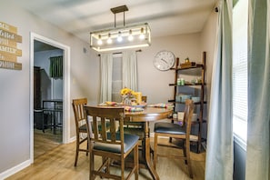 A dining area with a round table, wooden chairs, a chandelier, a wall clock, and a shelf with various items.