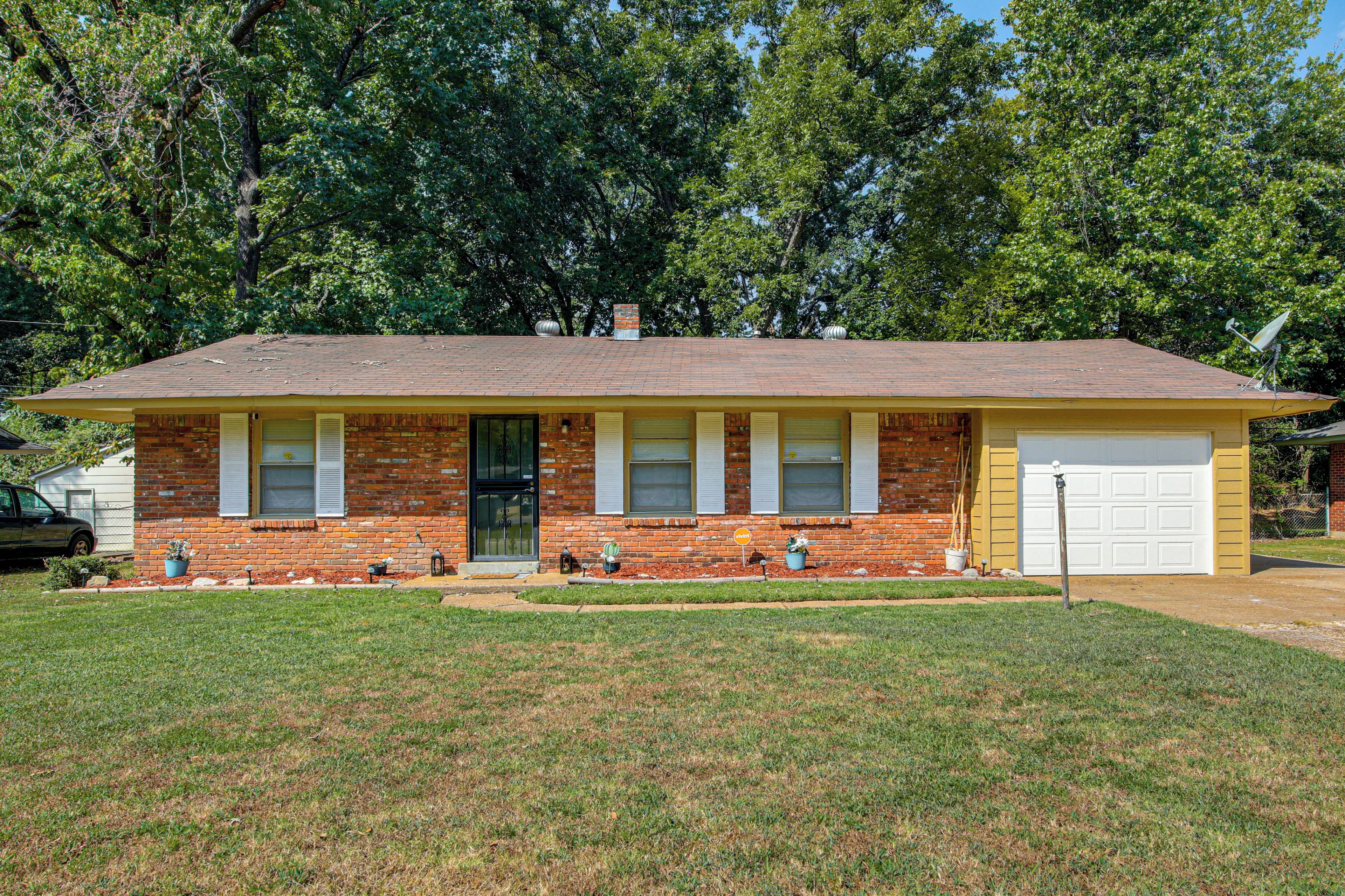A single-story brick house with a gabled roof, a front garage, and a small front yard with a lawn and potted plants.