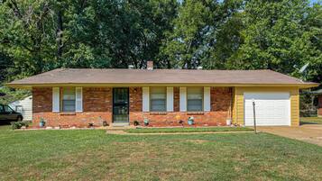A single-story brick house with a gabled roof, a front garage, and a small front yard with a lawn and potted plants.