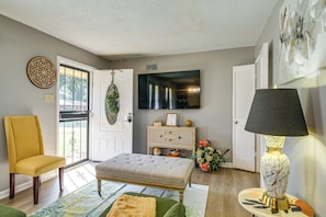A living room with a yellow armchair, a tufted ottoman, a patterned rug, a wall-mounted TV, and a lamp with a textured shade. - 4 Mi to Graceland! Cozy Home w/ Patio & Yard (Memphis)