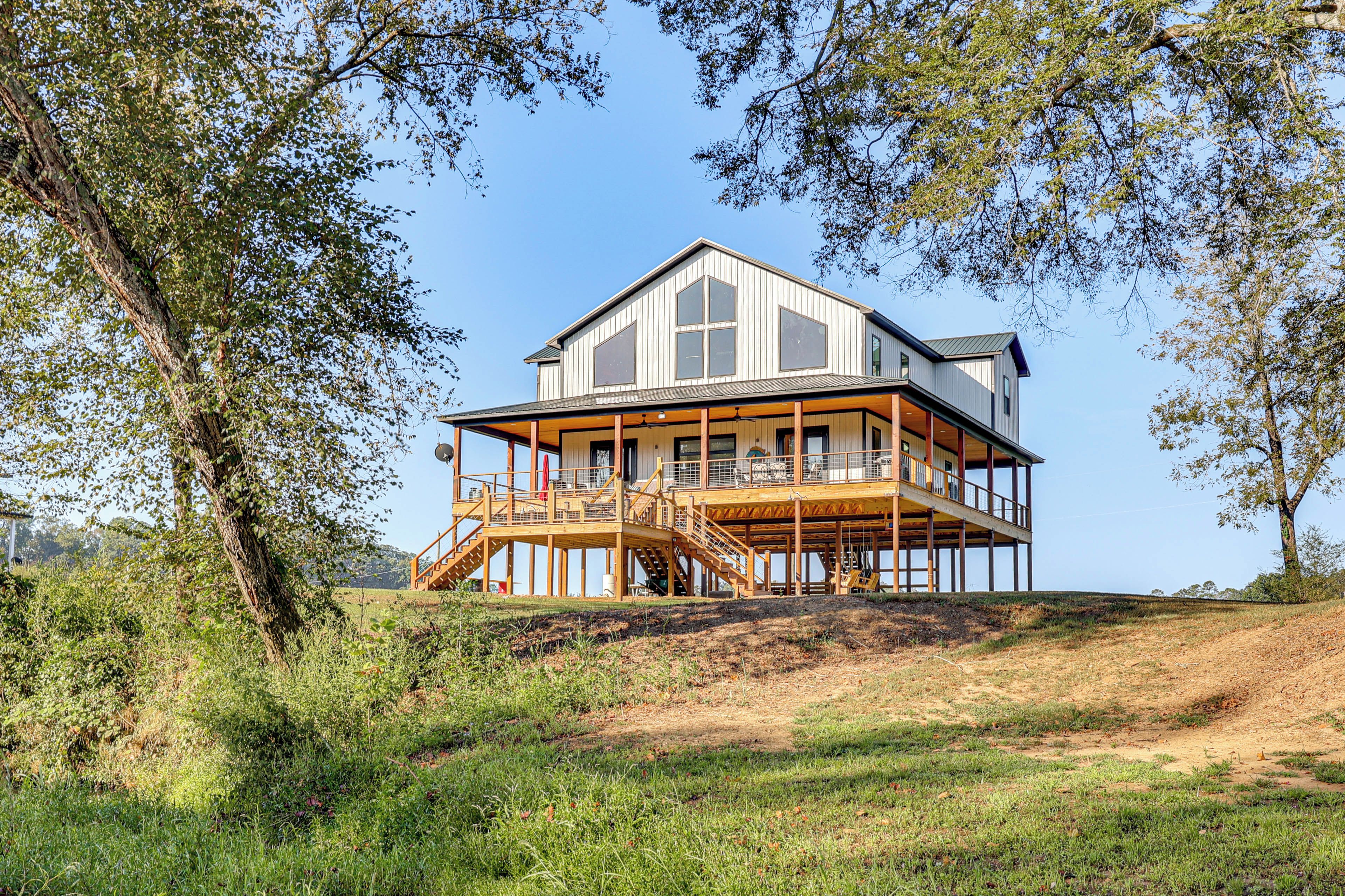 A two-story wooden house with a balcony, surrounded by trees and grassy area.