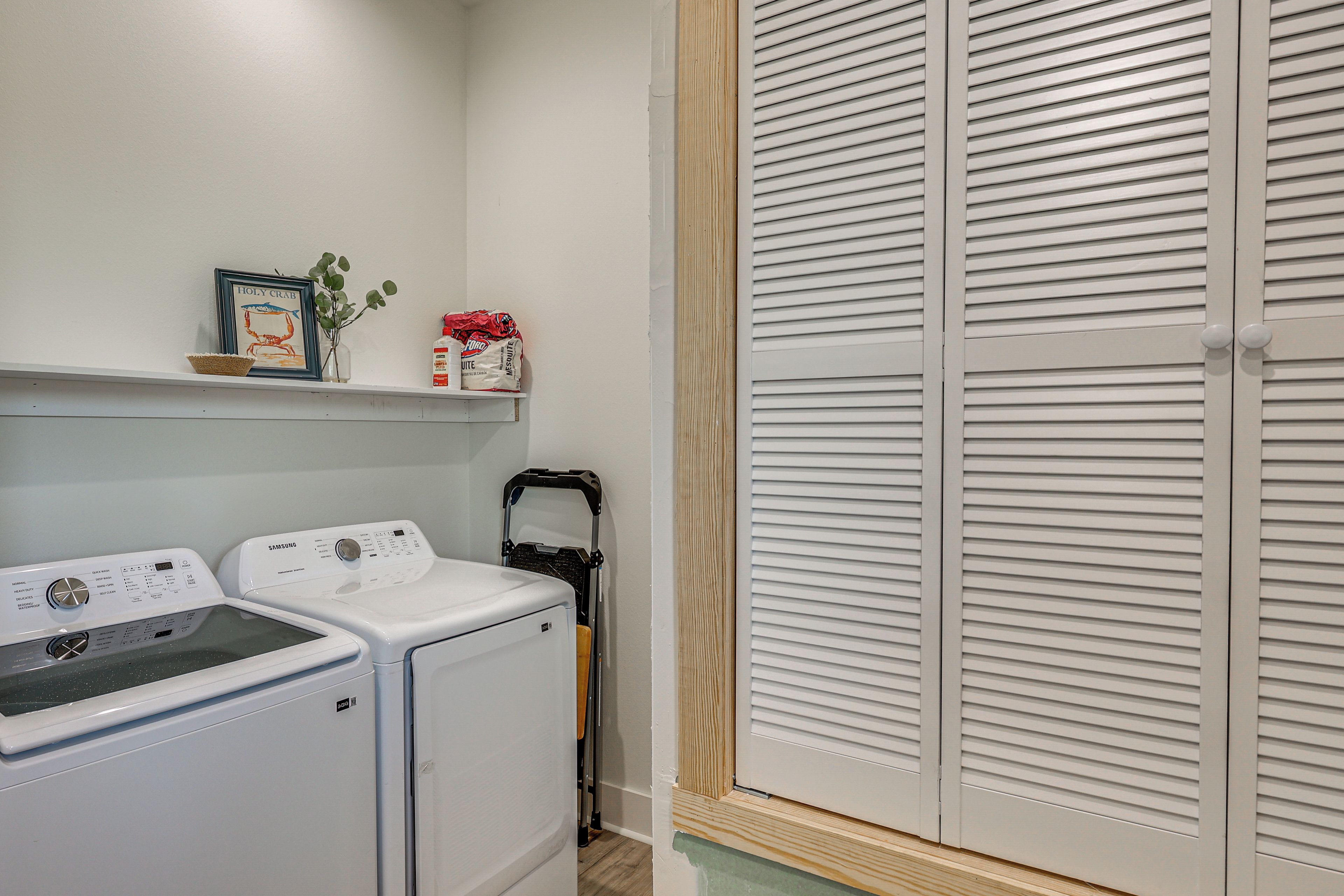 A laundry area with two washing machines, a shelf with a framed picture, and a closed closet with louvered doors.
