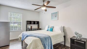 A bedroom with a bed, a desk, a ceiling fan, a window with blinds, and a wall-mounted shelf.