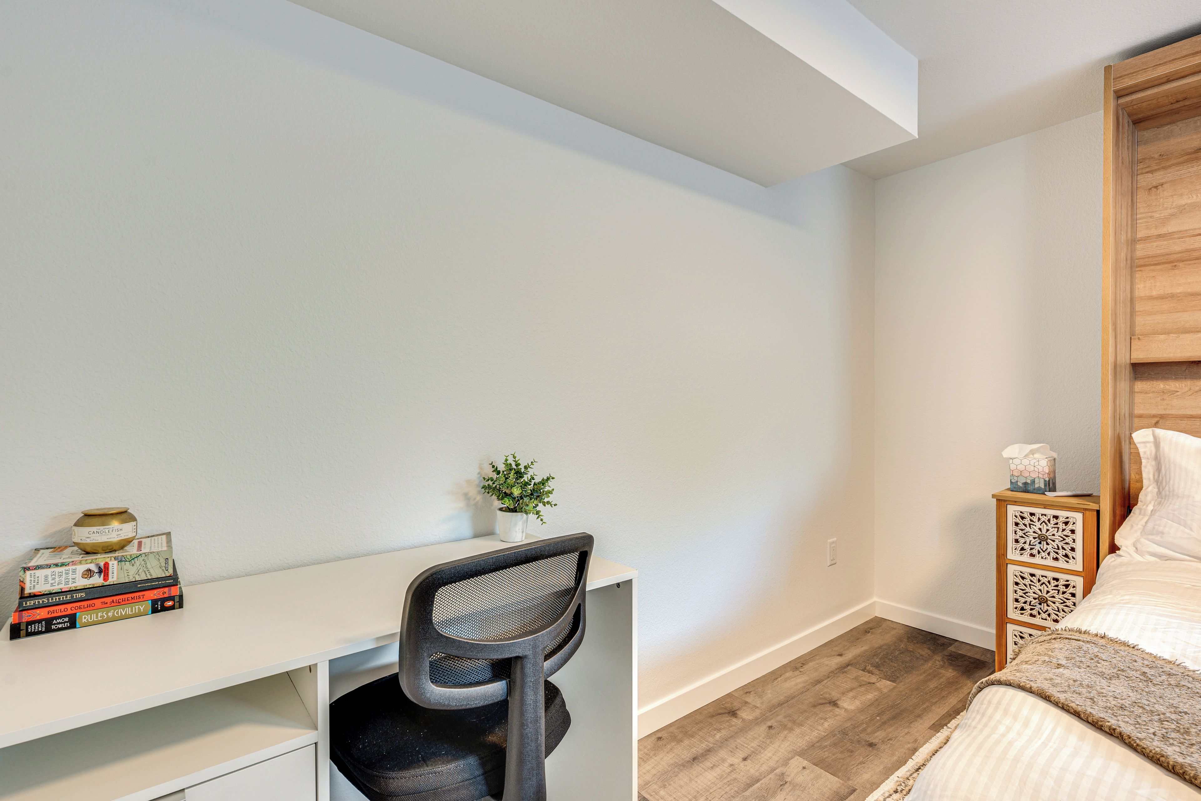 A bedroom with a wooden headboard, a white desk with a chair, a small plant, and a bedside table with a tissue box.