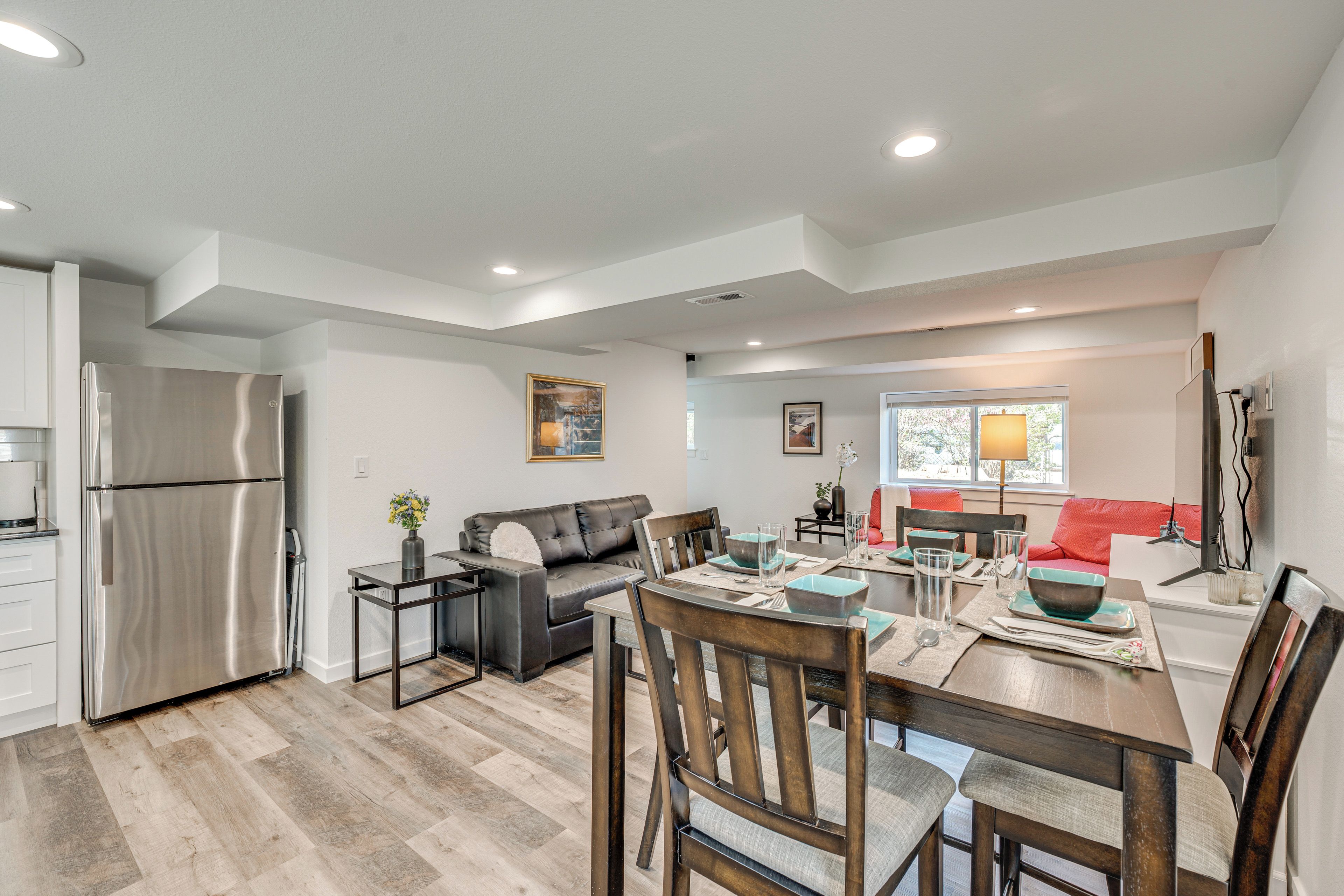 A modern kitchen with a stainless steel refrigerator, a dining area with a wooden table and chairs, a gray sofa, and a red couch.