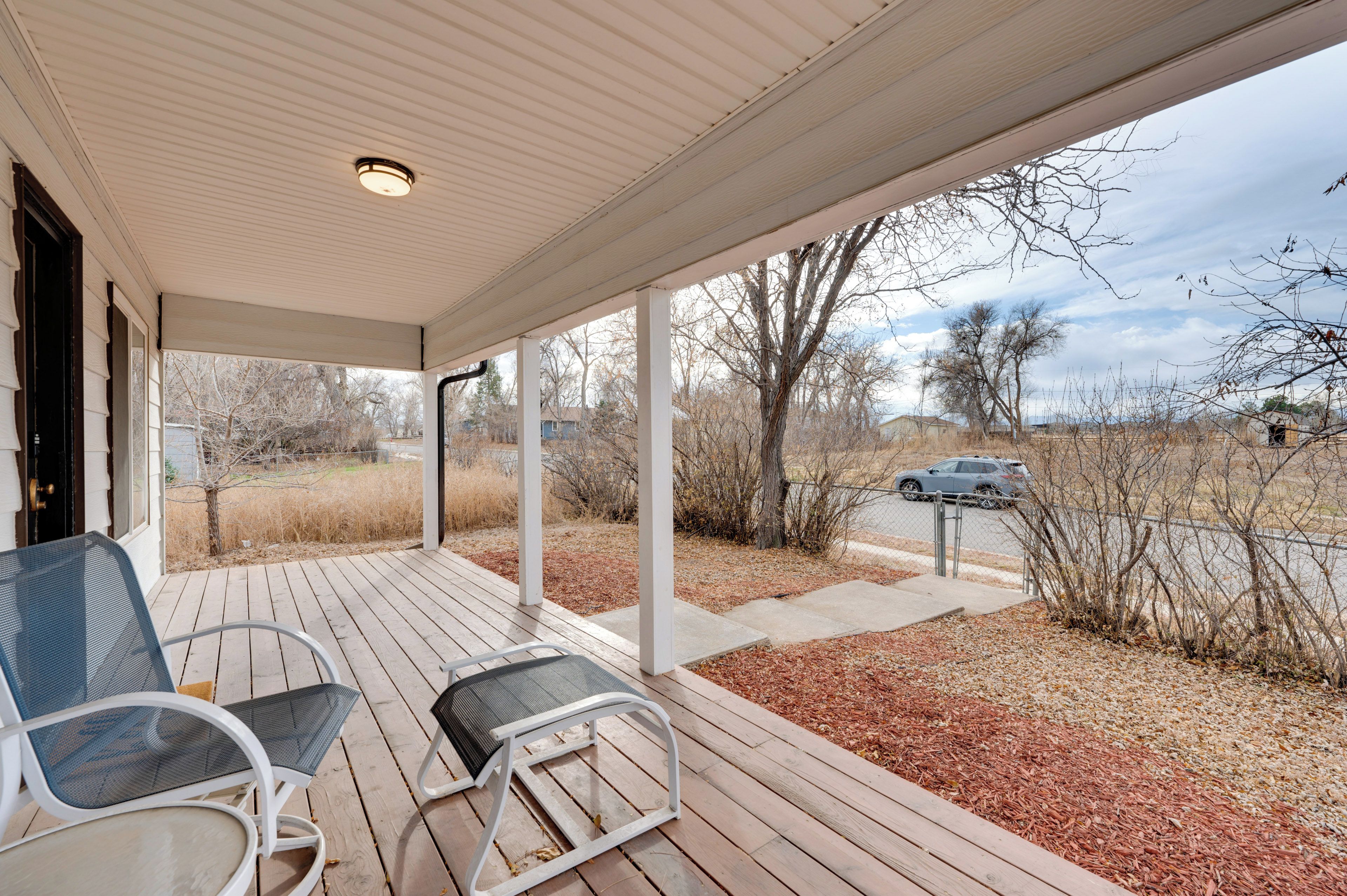 A covered porch with two chairs, a view of a road with a car, and a landscape with trees and shrubs.