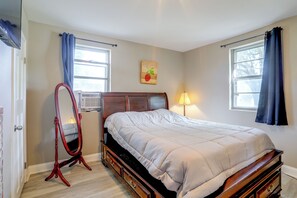 Quarto com cama, cômoda de madeira, espelho, radiador e duas janelas com cortinas.