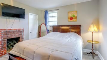 Un dormitorio con cama, televisor montado en la pared, chimenea, ventana con cortinas y lámpara.