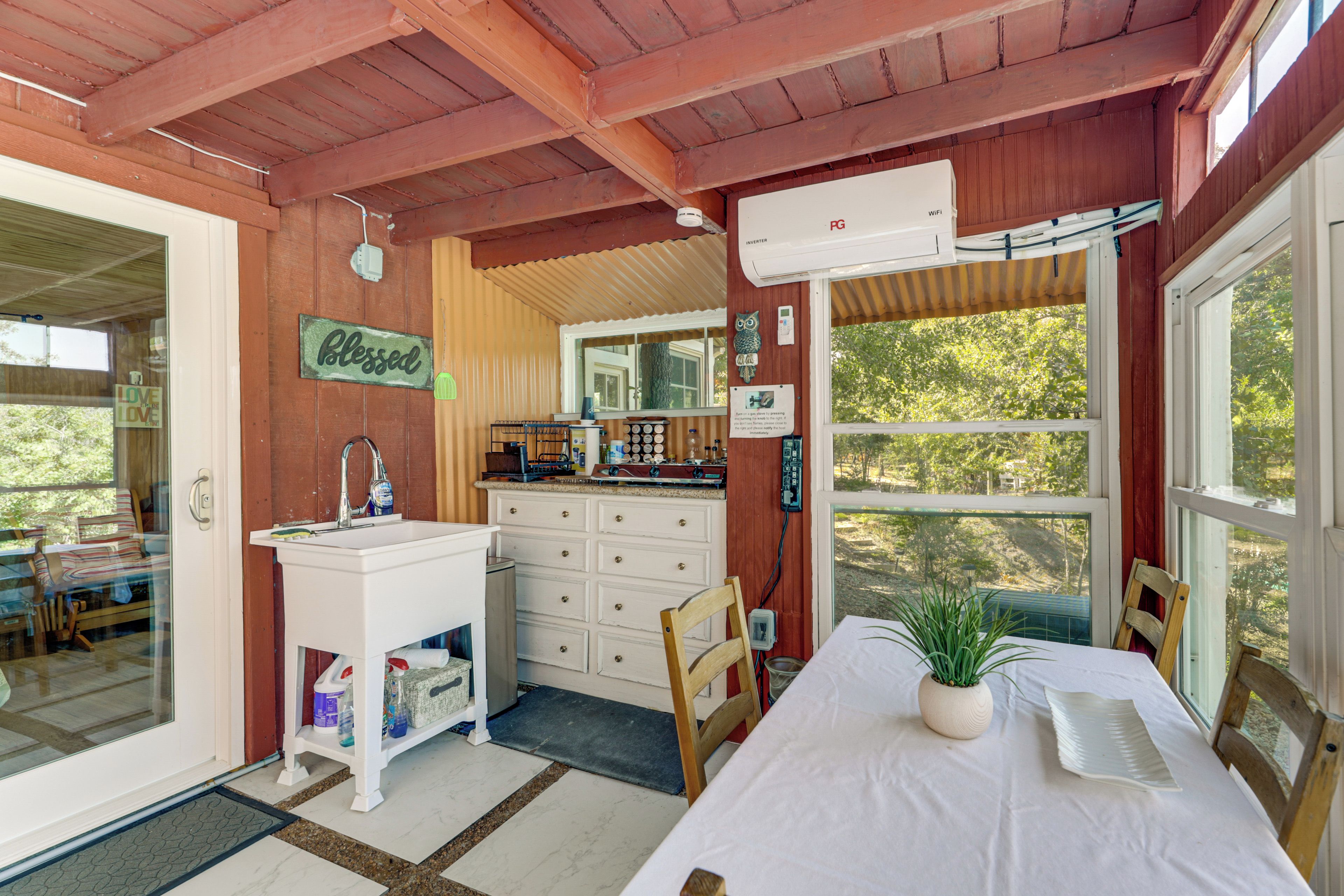 A cozy cabin interior with a wooden ceiling, a white sink, a dining table with chairs, and a large window offering a view of trees.