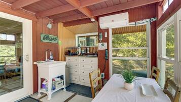 A cozy cabin interior with a wooden ceiling, a white sink, a dining table with chairs, and a large window offering a view of trees.