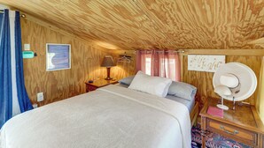 Quarto com teto de madeira, cama com roupa de cama branca, mesa de cabeceira com abajur, relógio de parede e um pôster.