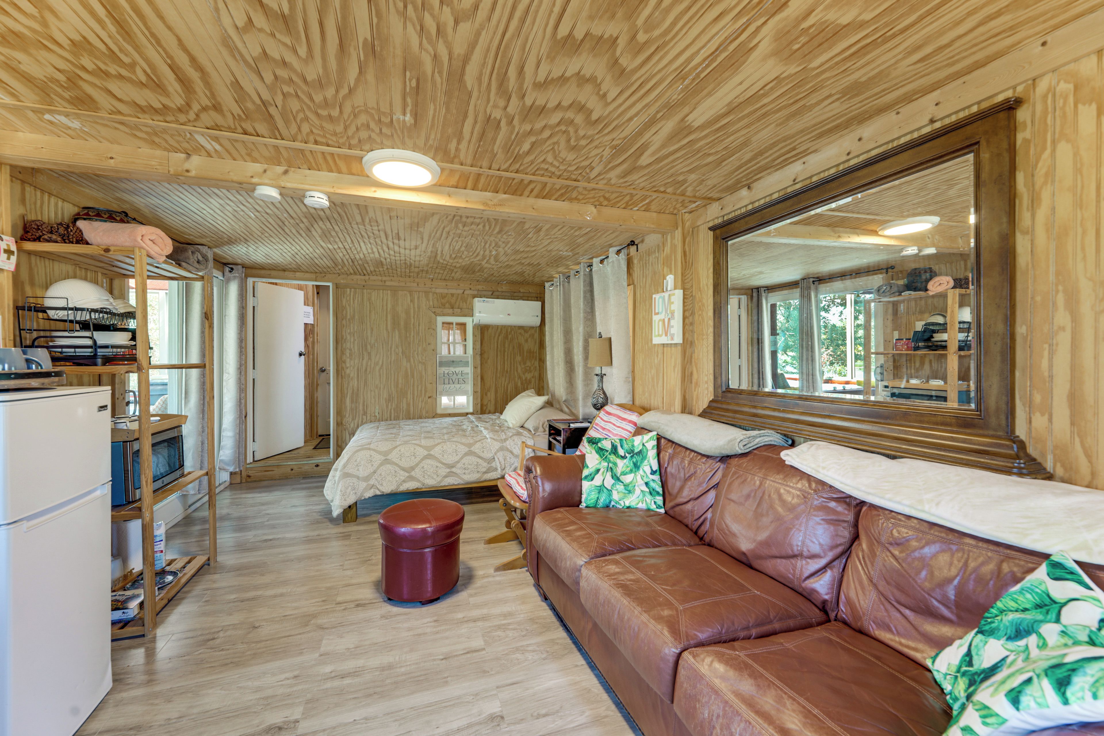 A cozy room with a wooden ceiling, a brown leather sofa, a small wooden stool, a bed with a patterned bedspread, and a window with curtains.