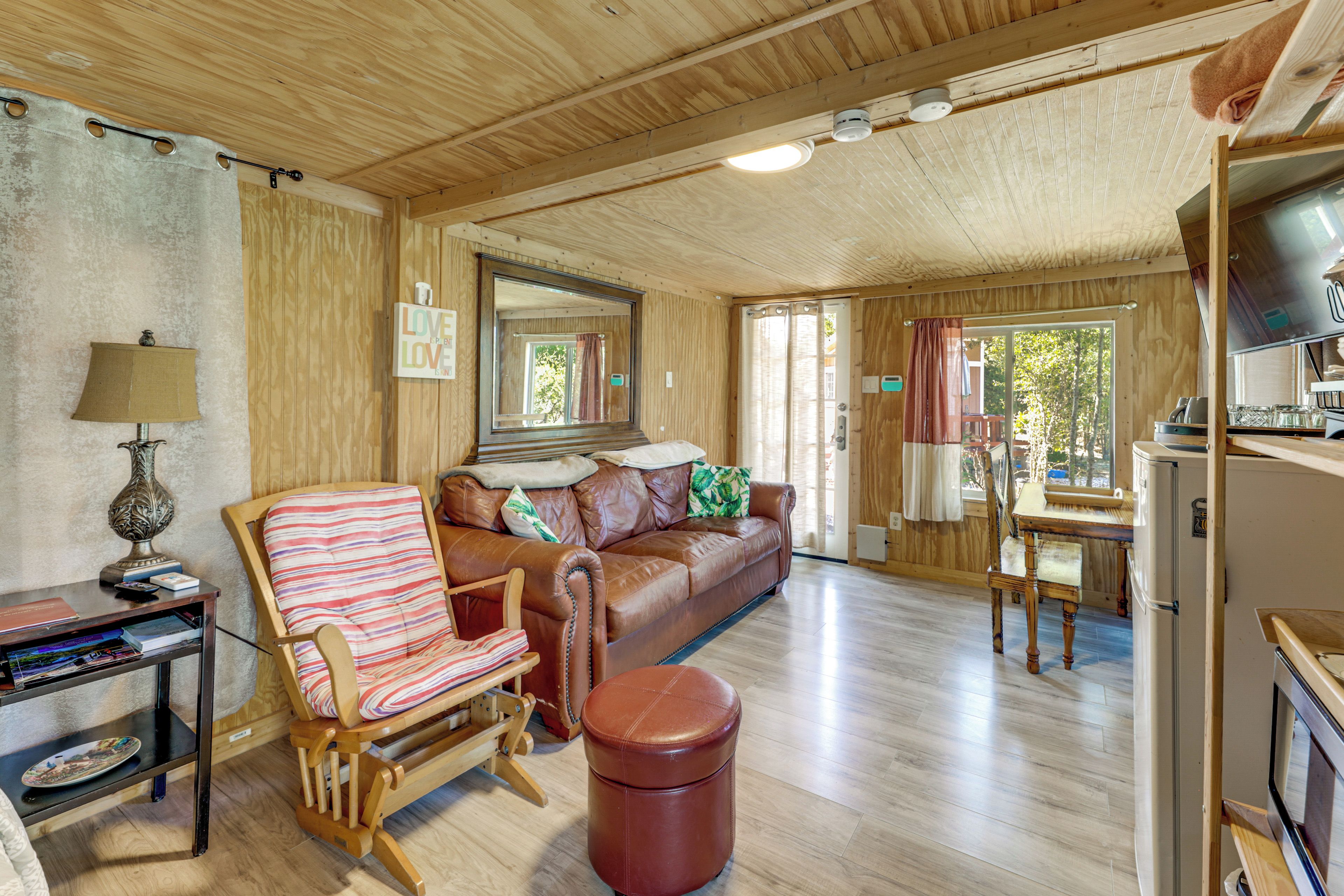 A cozy cabin interior with a wooden ceiling, a brown leather sofa, a striped armchair, a small table, and a television.