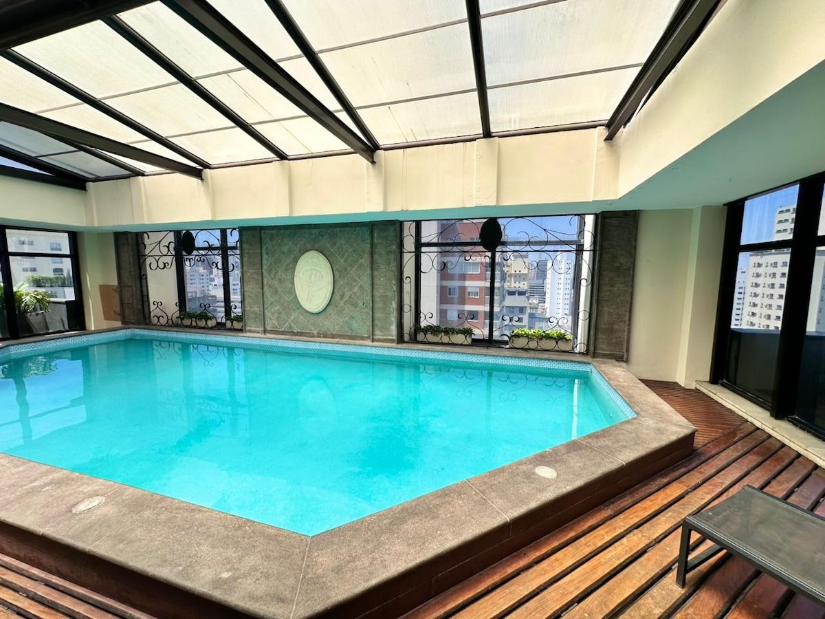 Une piscine intérieure avec un sol carrelé, entourée de fenêtres dotées de garde-corps en métal, et une zone de terrasse en bois.