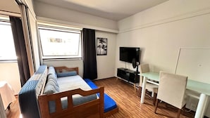 A compact room with a bed, a desk, a chair, a television, and a window. - Le Premier Suites (São Paulo)